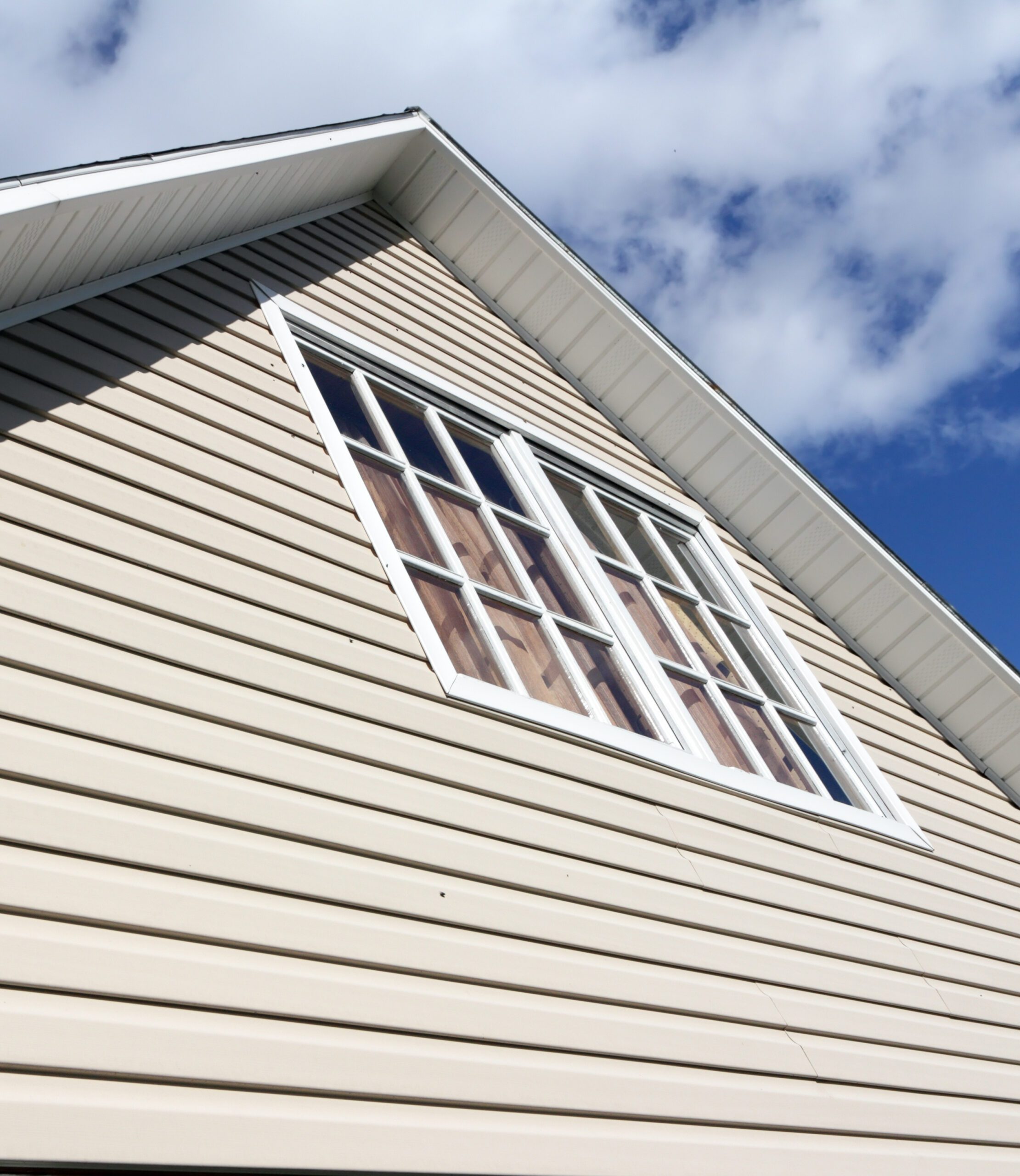 Close-up view of the upper side of a beige house with vinyl siding and a double window under a blue sky with clouds.